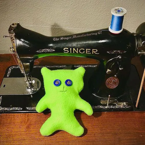 A photo of a green doll sitting next to an 83 year old pedal powered singer sewing machine.