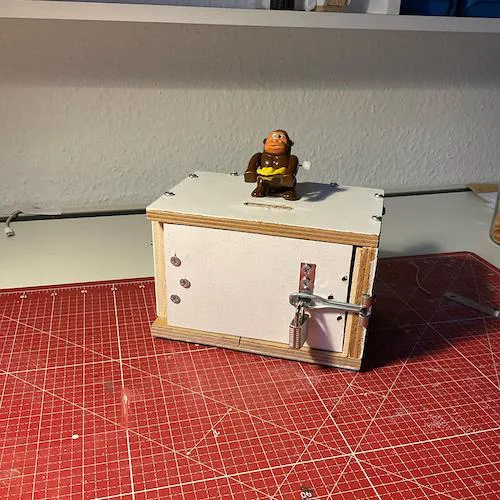 A photo of a white lock-box with a small wind up monkey toy glued on top.